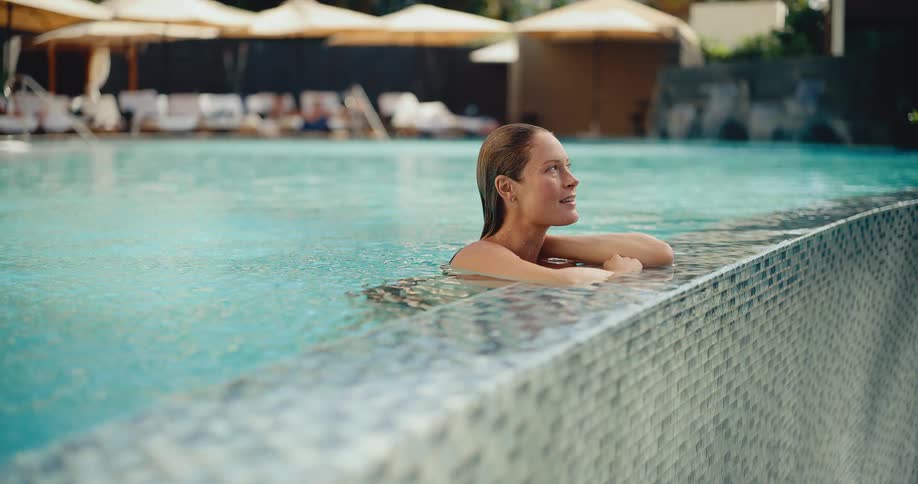 A happy woman with wet hair smiles and relaxes while leaning on the edge of a luxurious resort swimming pool