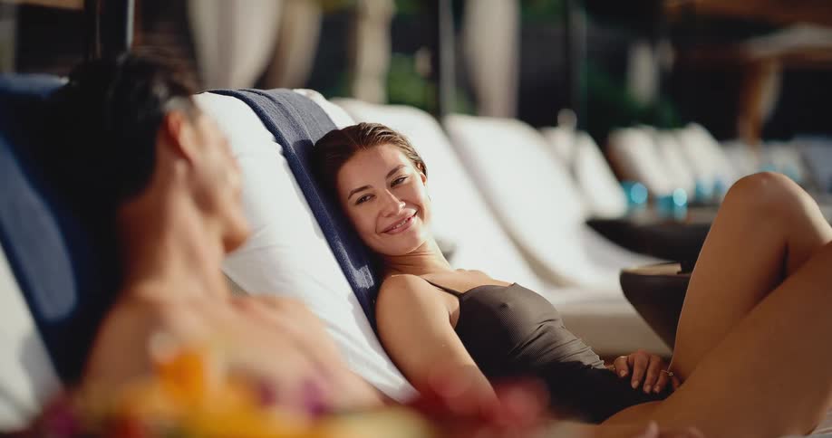 An attractive woman in a swimsuit smiles lovingly at her partner while relaxing together on lounge chairs at a luxury resort