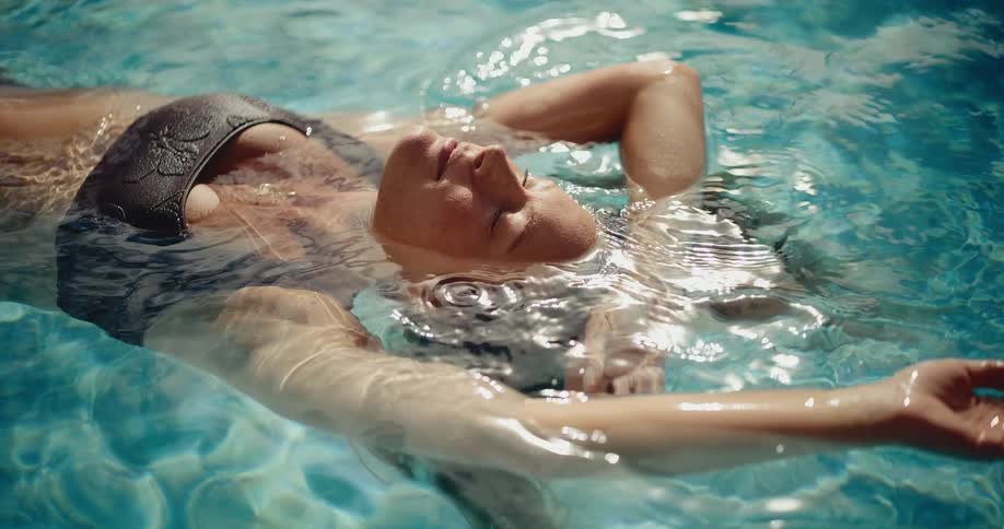 In slow motion, a woman in a black bikini relaxes as she floats on her back in the sparkling clear water of a swimming pool