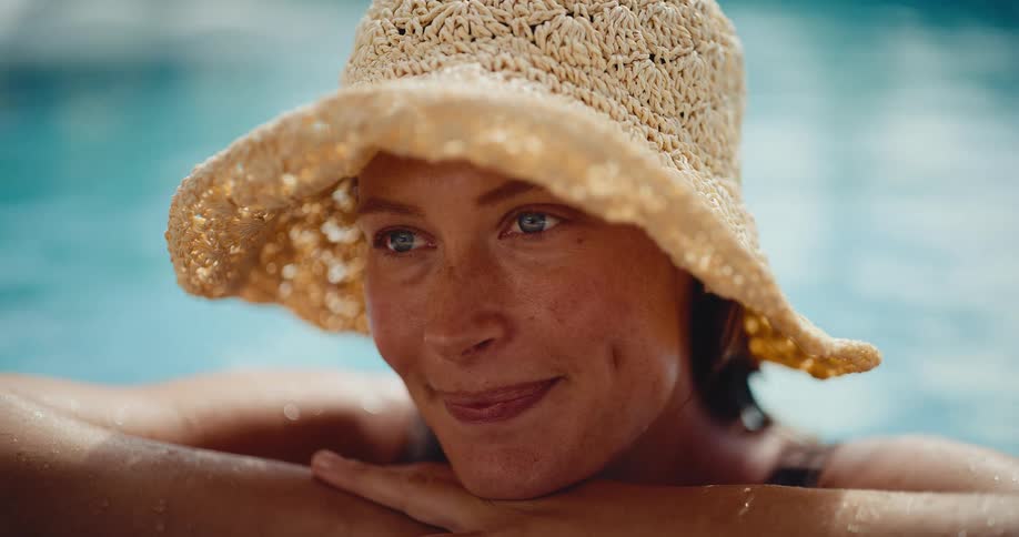 A beautiful woman in a straw hat smiles and relaxes in a sunlit swimming pool, enjoying a peaceful summer day