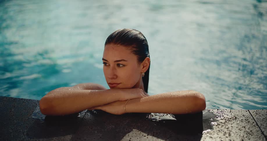 Close-up portrait of a beautiful young woman with wet hair relaxing on the edge of a swimming pool, gazing calmly at the camera