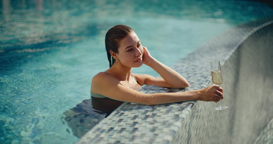 A serene young woman with wet hair relaxes at the edge of a sunlit swimming pool with a glass of champagne
