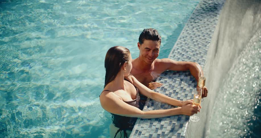 A smiling couple relaxes in a luxury swimming pool, talking and toasting with glasses of champagne on a sunny day