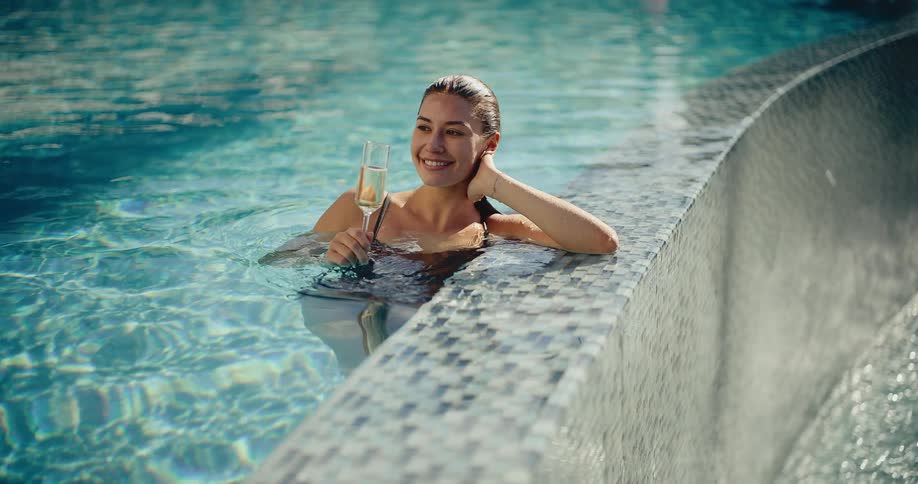 A beautiful woman in a swimsuit relaxes at the edge of a luxurious swimming pool, enjoying a glass of champagne on a sunny day