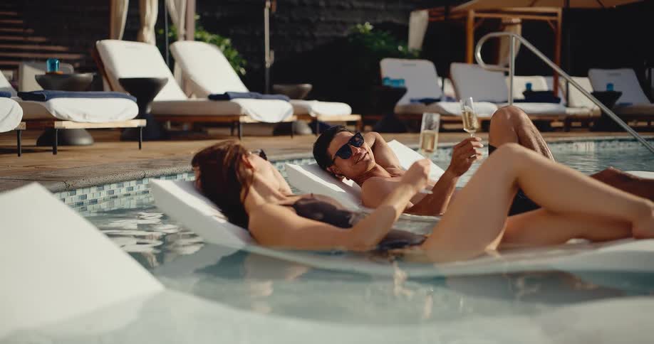 A young couple relaxes on in-water lounge chairs, toasting with champagne at a luxury resort pool