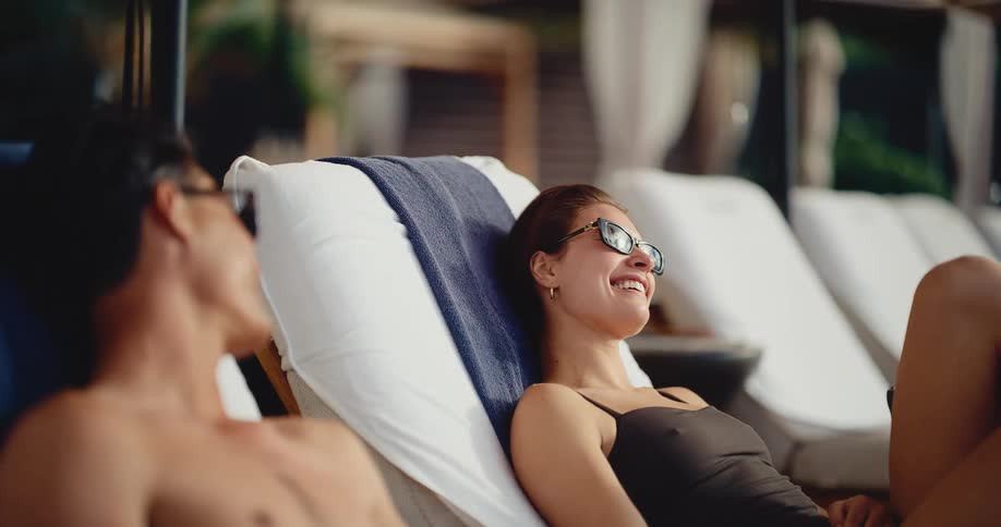 A beautiful woman in sunglasses smiles warmly at her partner as they relax together on lounge chairs at a luxurious poolside resort