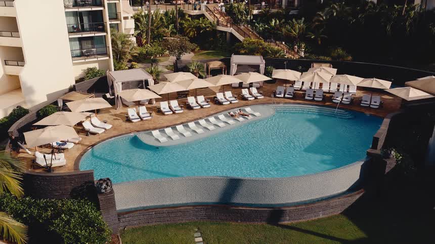 High-angle time-lapse of a luxurious resort swimming pool with lounge chairs and umbrellas as shadows move across the scene