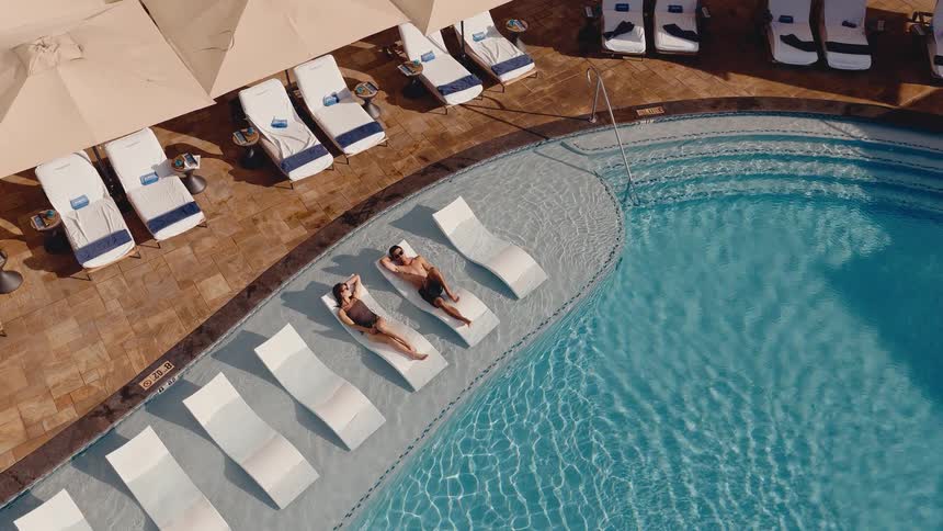 An aerial view shows a couple relaxing on modern lounge chairs submerged in the shallow end of a beautiful resort swimming pool