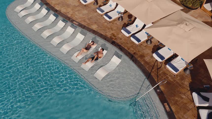 A young couple relaxes on in-water chaise lounges at a luxury resort pool in this aerial top-down shot that slowly pulls away