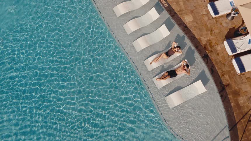 An aerial top-down view shows a couple relaxing and sunbathing on lounge chairs in a sparkling blue swimming pool at a luxury resort