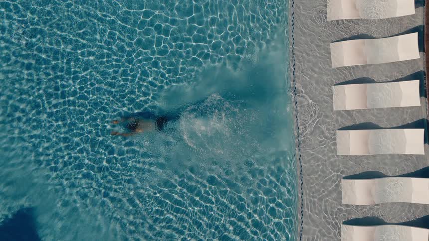 Slow-motion aerial top-down view of a man swimming breaststroke in a clear blue luxury swimming pool next to white lounge chairs