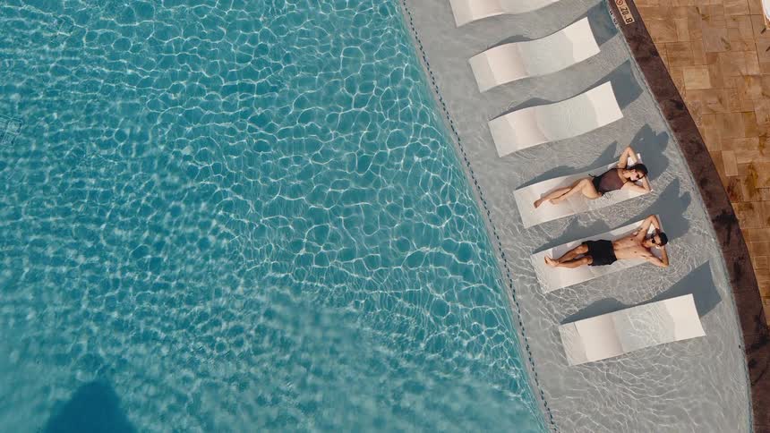 Aerial top-down view of a couple sunbathing on lounge chairs in the shallow end of a luxury swimming pool