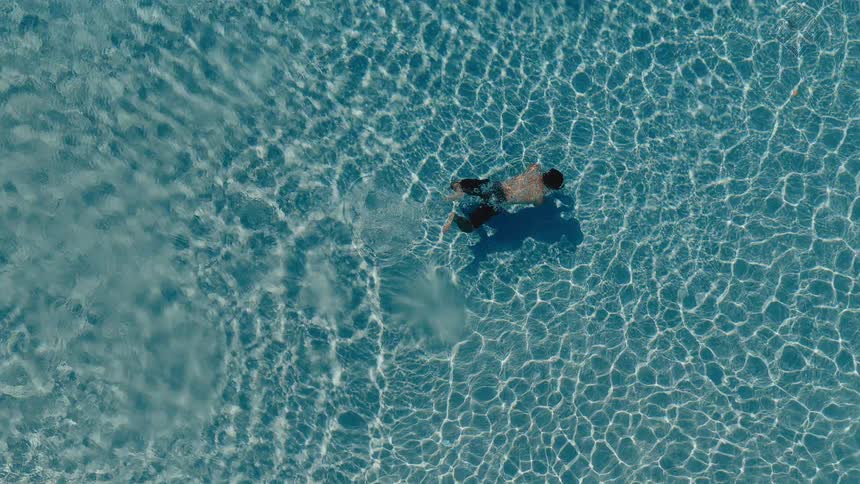 Top-down aerial view of a man gracefully swimming in slow motion across a clear blue swimming pool