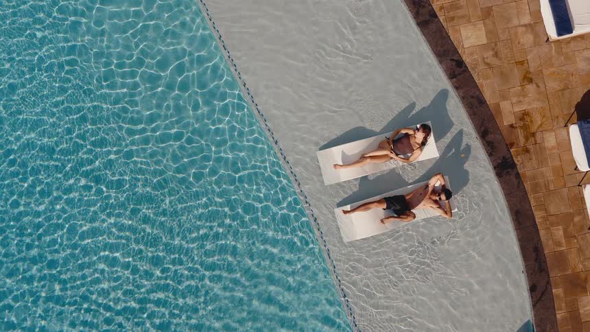 A slow aerial top-down shot of a couple relaxing on pool loungers in the shallow end of a resort swimming pool