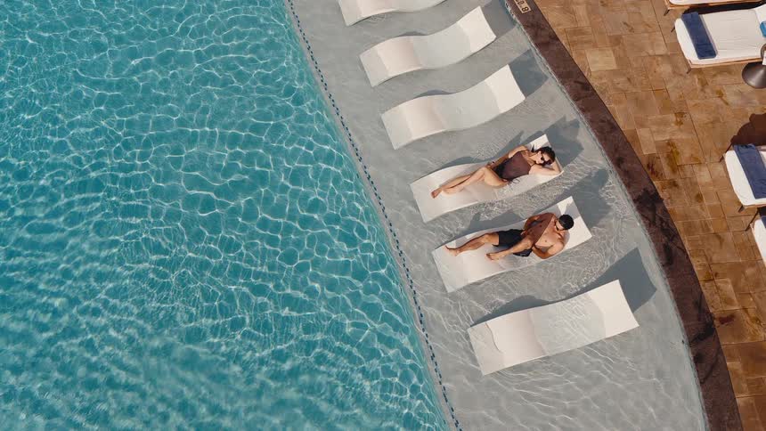 Top-down aerial view of a couple relaxing on in-water chaise longues at a luxury resort pool