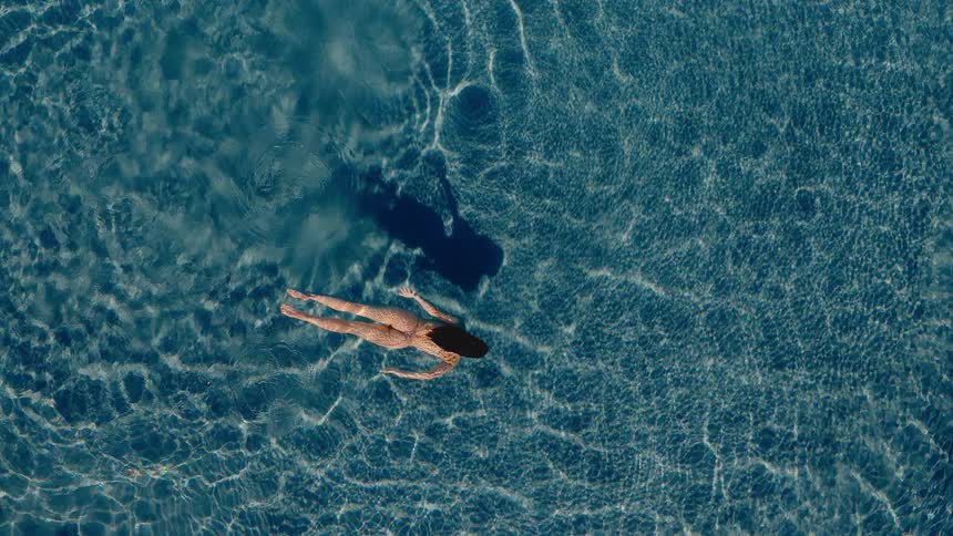 A tranquil top-down aerial view of a woman in a bikini floating serenely in a sun-dappled, clear blue swimming pool