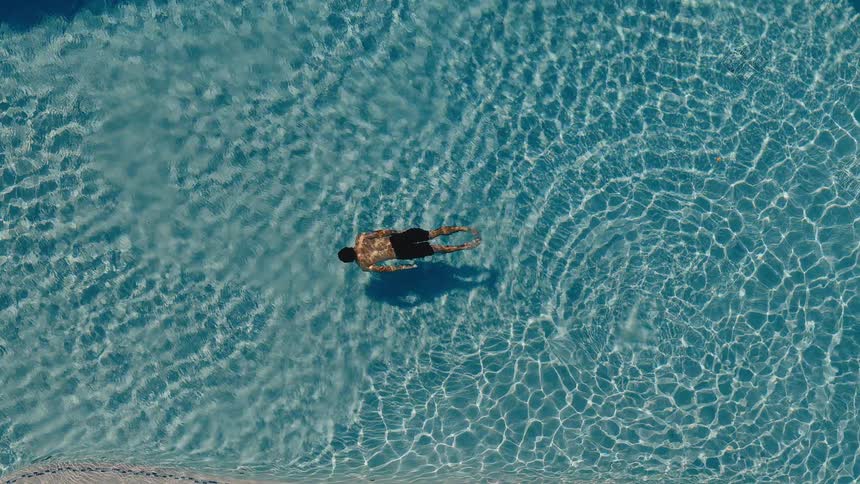 An aerial top-down view of a man floating on his back and relaxing in a crystal clear blue swimming pool on a sunny day