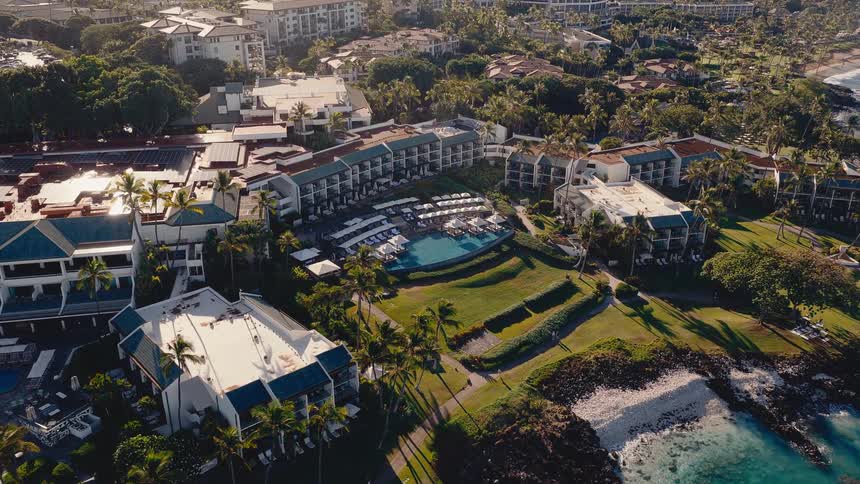 Drone slowly flies towards a luxurious Hawaiian resort, revealing a beautiful swimming pool and the stunning turquoise ocean coastline