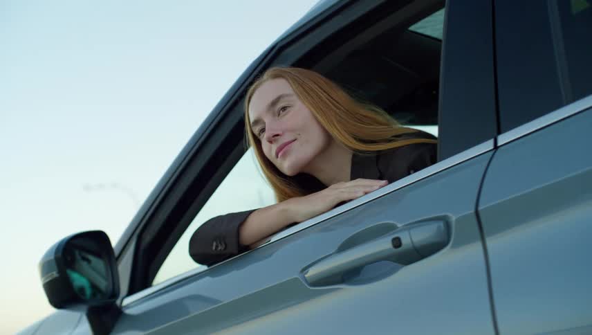 A happy young woman with long red hair looks out of the driver's side window of her car with a gentle smile