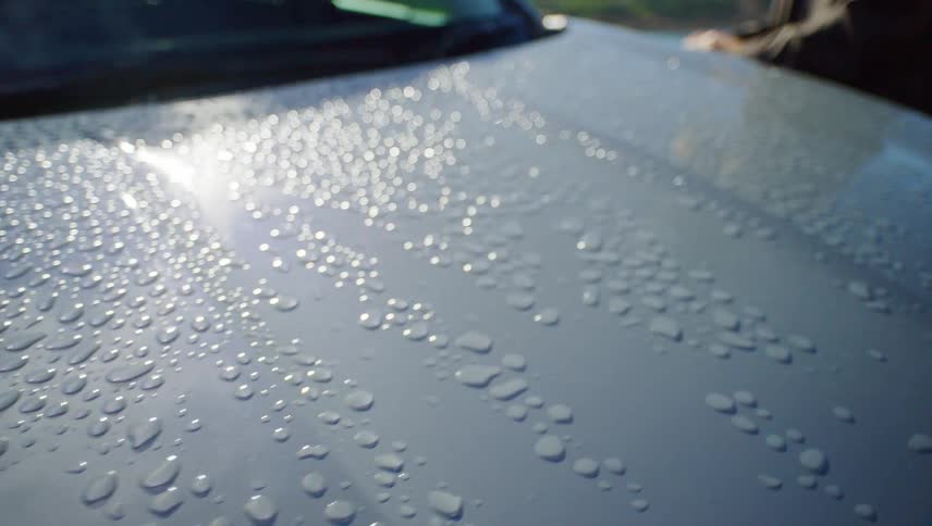 Slow motion close-up of water droplets beading on the hood of a car, glistening in the bright sunlight