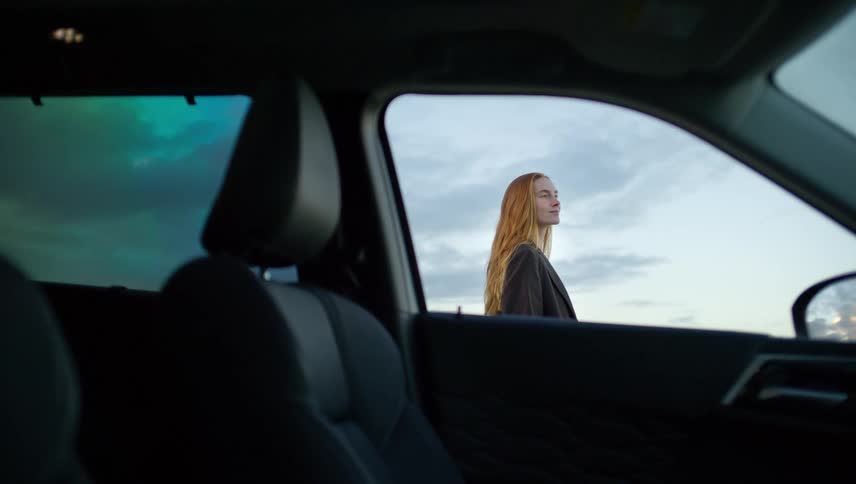Interior car view of a young woman with long red hair standing outside and looking at the scenic sunset