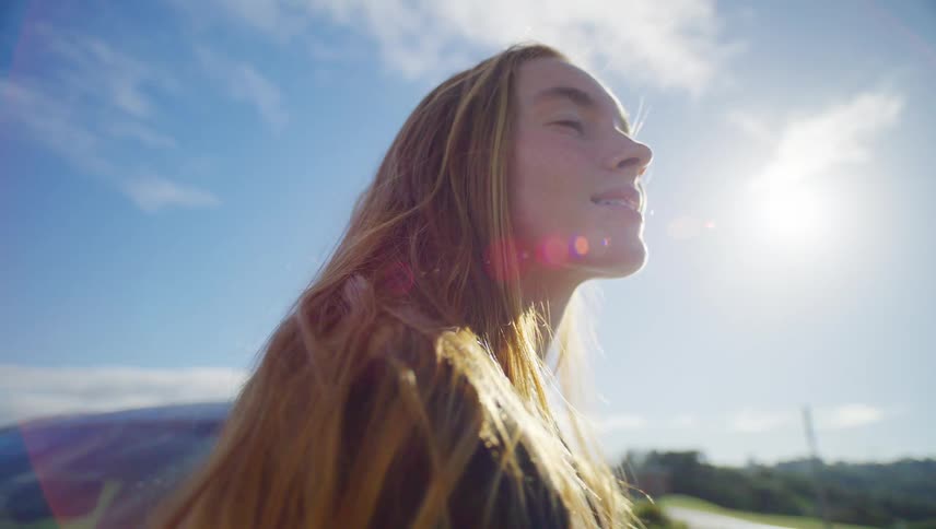 Slow motion close-up of a contemplative young woman with windblown hair looking up at the bright sun