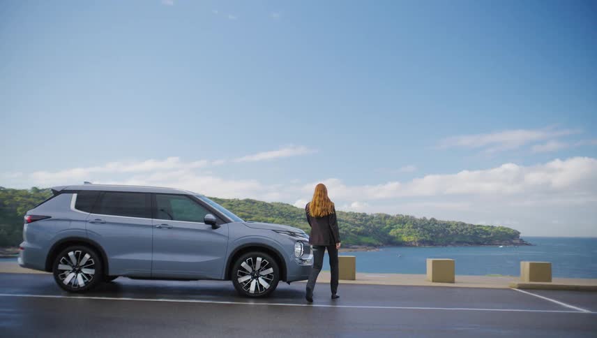 Slow motion side view of a well-dressed woman getting out of her modern SUV, parked on a coastal road, to admire the beautiful ocean view