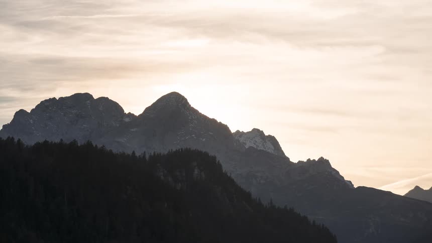 A beautiful time-lapse of the sun setting behind a majestic mountain range, silhouetting the peaks against a soft, glowing sky