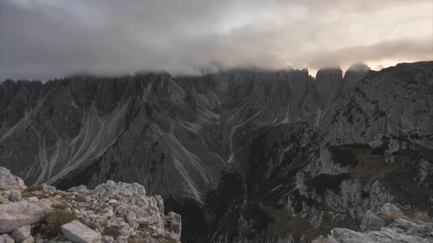 A moody time-lapse shows clouds and fog rolling over a dramatic and rugged mountain range as the camera slowly zooms out