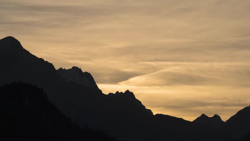 A golden hour timelapse shows clouds and contrails drifting over a silhouetted mountain range