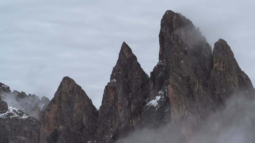 A dramatic time-lapse shows thick clouds and fog swirling around the jagged, snow-dusted peaks of a mountain range