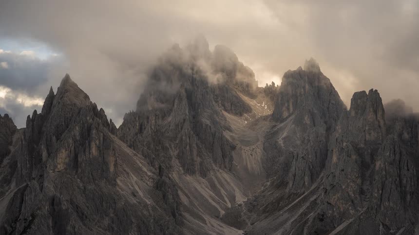A moody time-lapse of dramatic clouds swirling around the jagged peaks of a mountain range at sunset