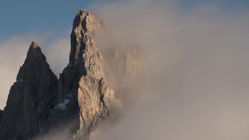 Slow-moving clouds drift across a rugged mountain peak, gradually revealing its sunlit, rocky face against a blue sky