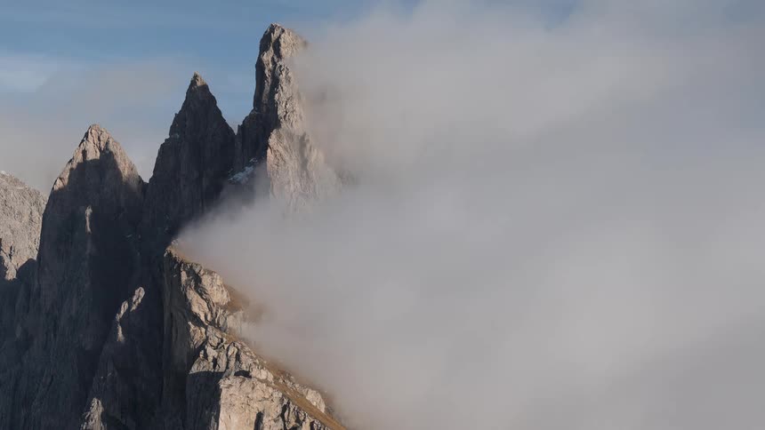 A thick bank of fog slowly rolls past, unveiling the sharp, rocky peaks of a mountain range under a blue sky
