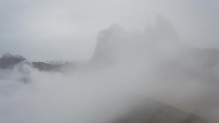 A dramatic aerial view of clouds and fog rolling over the majestic, snow-capped peaks of the Seceda ridgeline in the Dolomites
