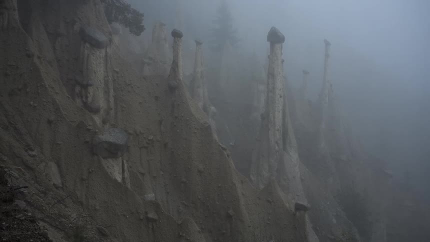 Mysterious tall earth pyramids, known as hoodoos, emerge from the dense fog on a steep mountainside