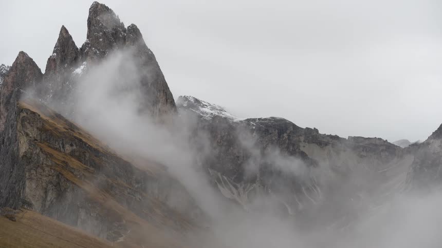 Thick, fast-moving fog and clouds swirl around the jagged peaks of a dramatic mountain range