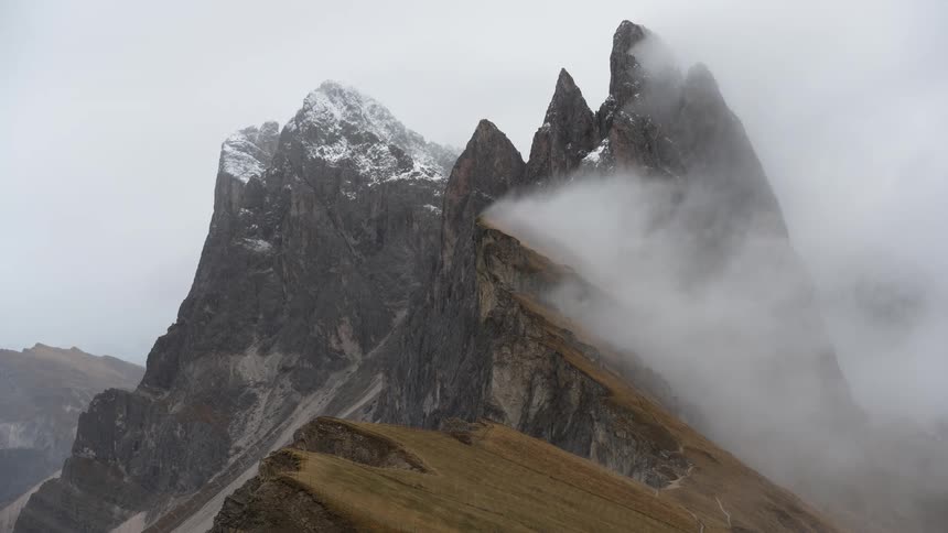 Thick fog slowly clears to reveal the jagged, snow-dusted peaks of a dramatic mountain range