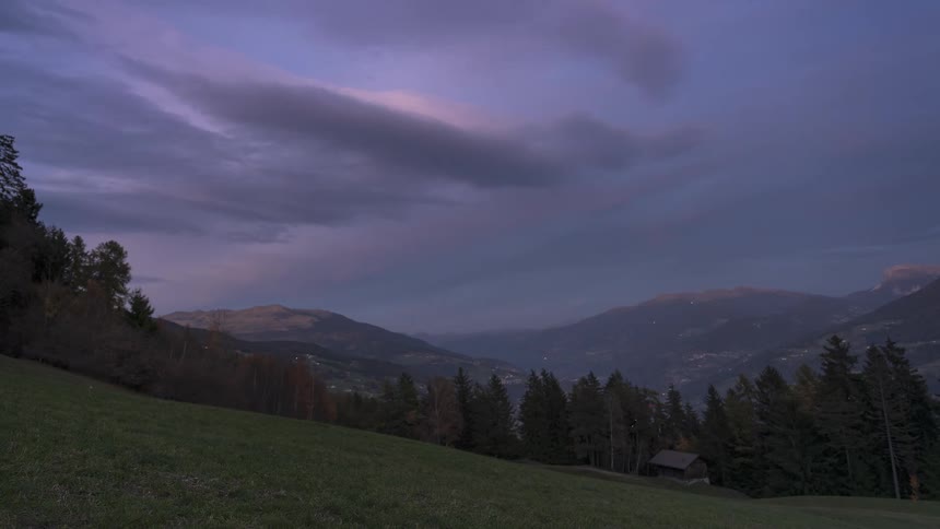 A scenic timelapse shows a colorful purple sunset transitioning to night over a vast mountain valley, as city lights begin to twinkle in the dark