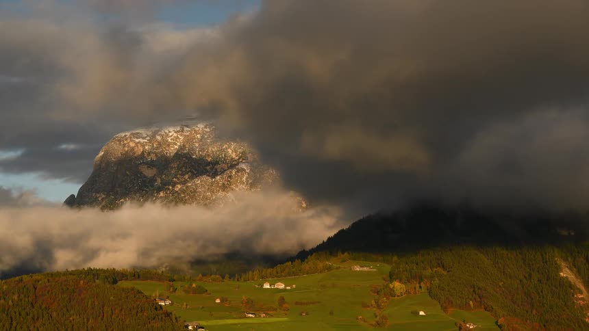A breathtaking aerial view shows a snow-dusted mountain peak emerging through shifting clouds at golden hour over a lush green valley