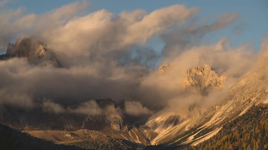 Clouds gracefully drift through a majestic mountain range, illuminated by the warm, golden light of sunset