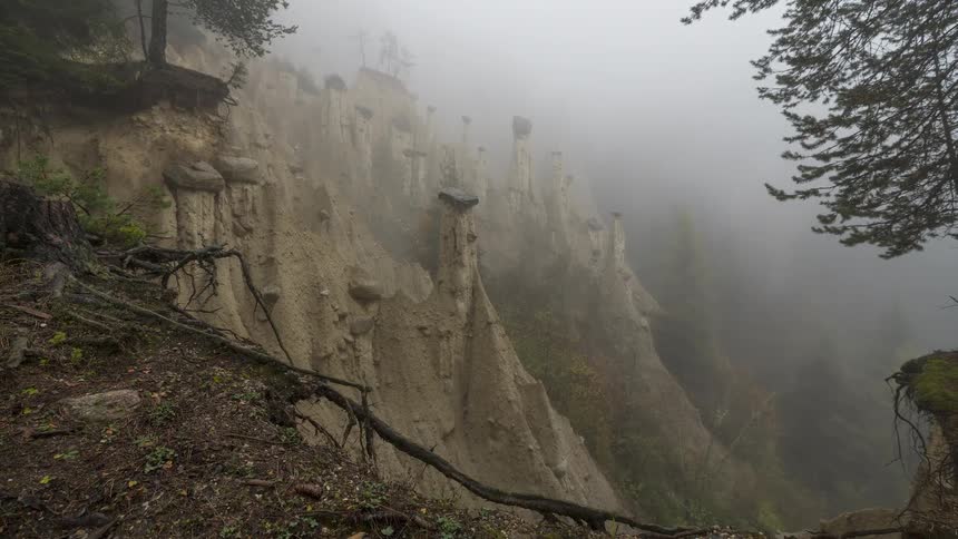 A spooky timelapse captures thick fog rolling through a forest, revealing unique earth pyramid rock formations on a steep hillside