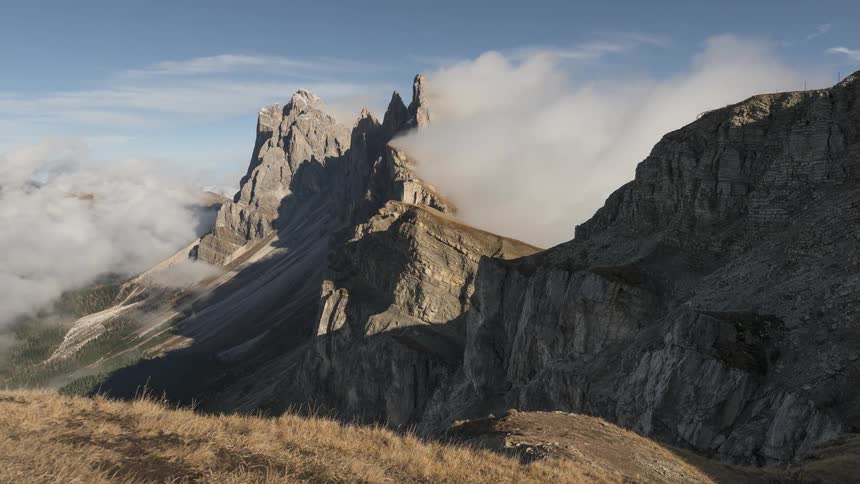 A stunning view of clouds swirling around the dramatic, jagged peaks of the Seceda mountain range in the Italian Dolomites