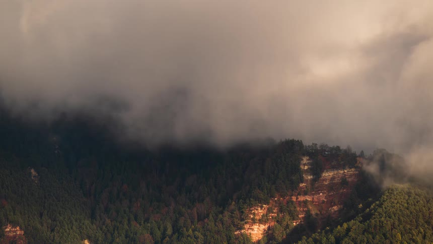 A time-lapse of dramatic clouds and fog rolling over a sunlit mountain forest, casting moving shadows across the landscape
