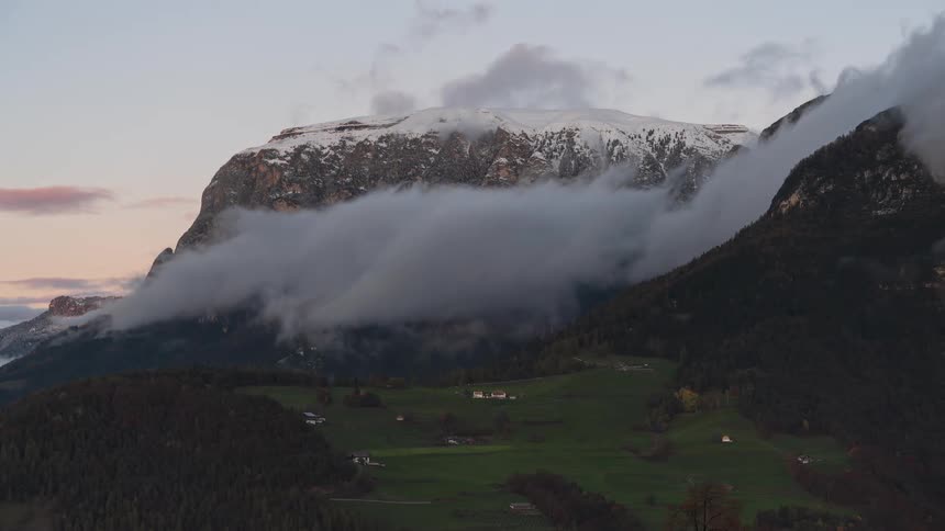 A dramatic time-lapse captures clouds rolling over a snow-capped mountain as the golden sunset light fades to twilight above a green alpine valley