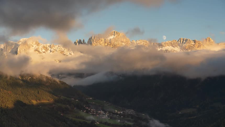 Golden hour light illuminates the snowy peaks of a mountain range as clouds drift over a valley with a quaint village, while the moon rises in the sky