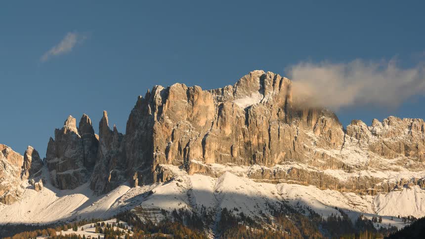 A majestic timelapse captures clouds flowing over the sunlit, snow-dusted peaks of a rugged mountain range against a clear blue sky