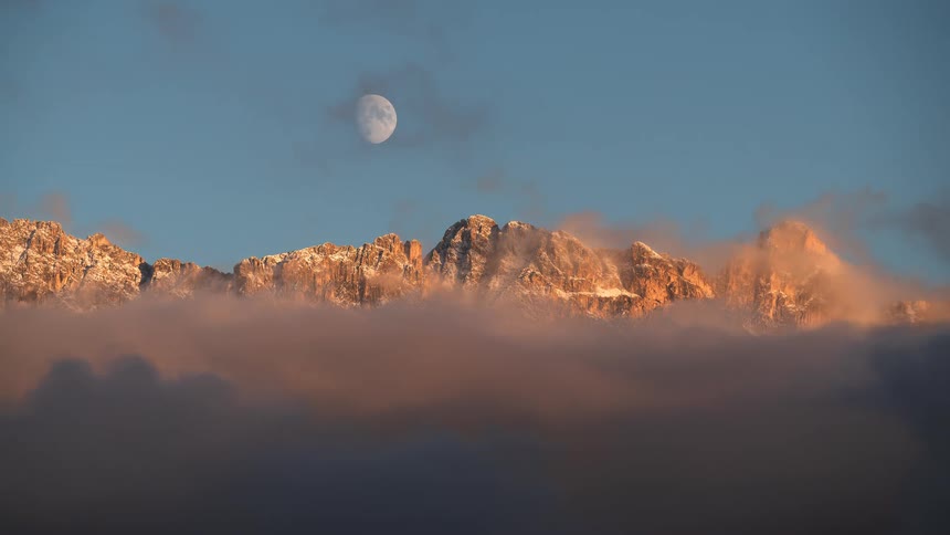 Timelapse of a snow-capped mountain range and moon at sunset as golden clouds roll in and obscure the view
