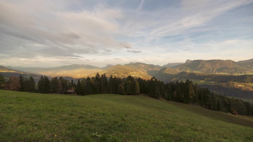 Golden sunlight illuminates a majestic mountain valley, viewed from a tranquil green meadow overlooking a dense pine forest