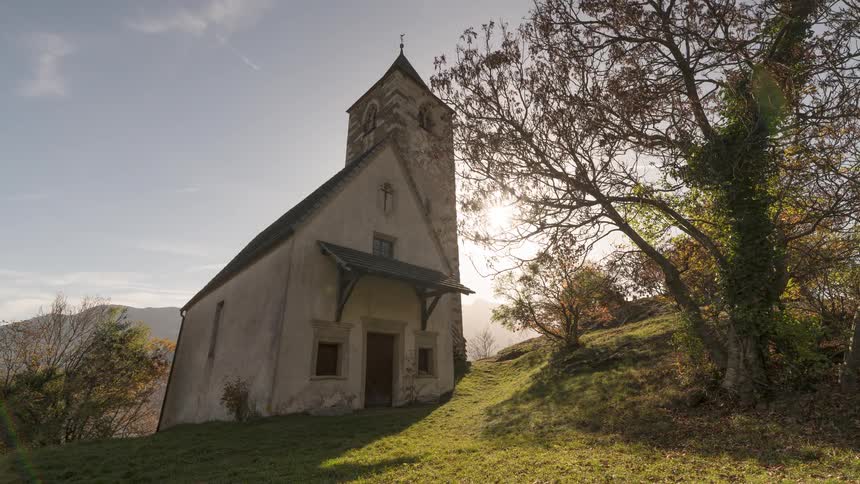 A peaceful timelapse shows the sun setting behind a rustic old church on a green hillside during autumn